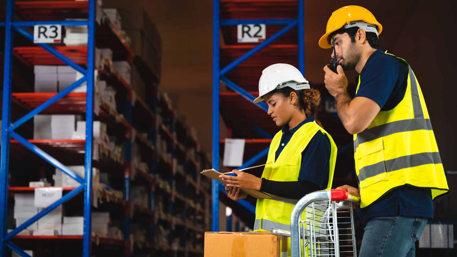Two warehouse workers in hard hats and hi-vis, one writing on a clipboard, the other on a radio