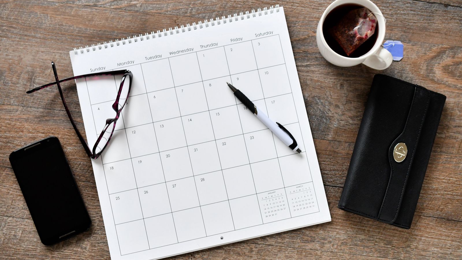 Blank wall calendar with a pen, glasses, a cup of tea, a phone and a wallet on a wooden desk