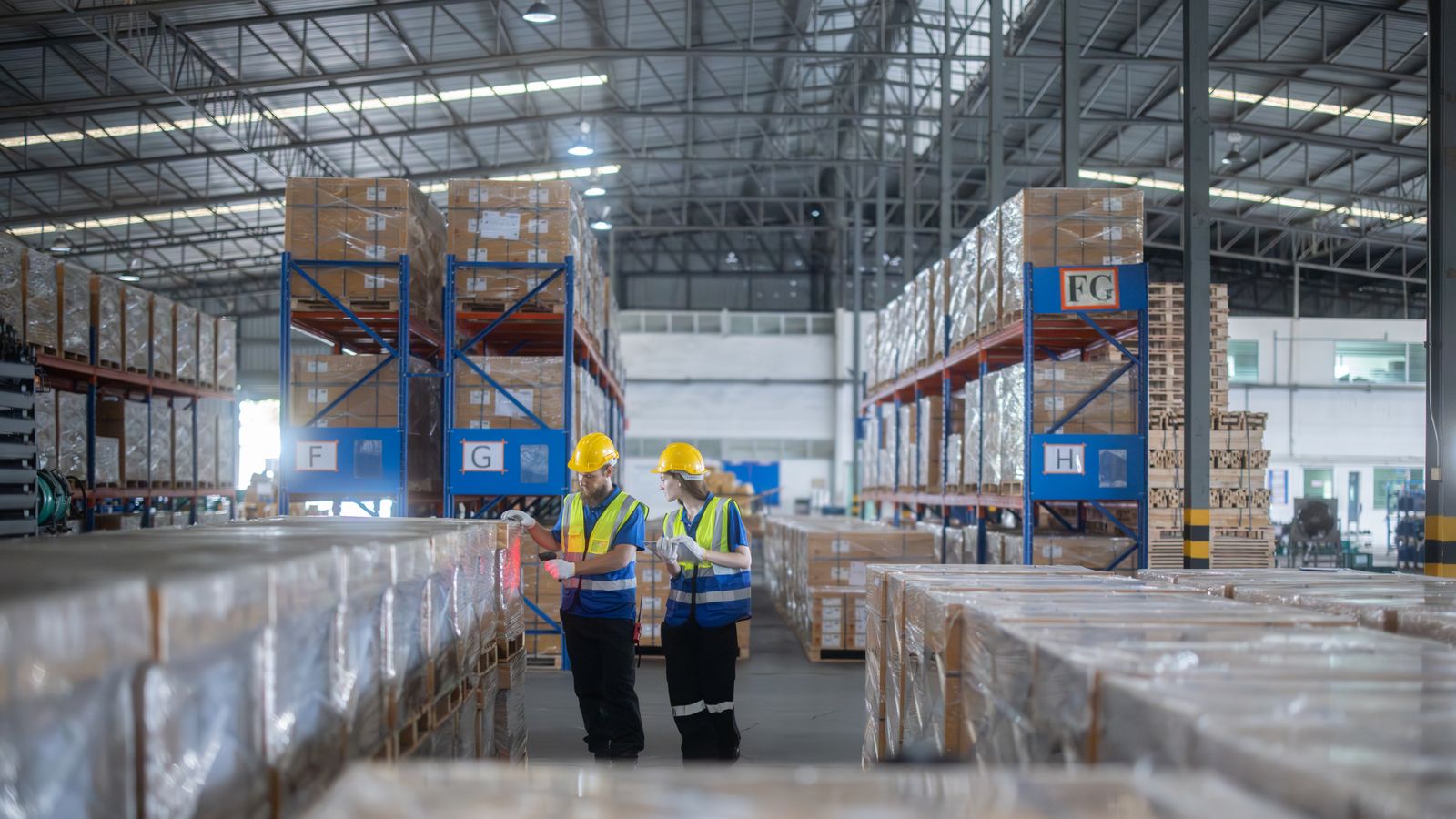 Two workers in hard hats and hi-vis vests checking stock between tall racks in a large warehouse