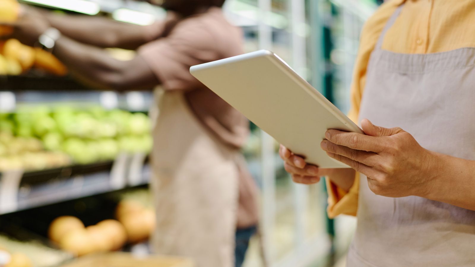 Shop worker in an apron holding a tablet beside produce shelves