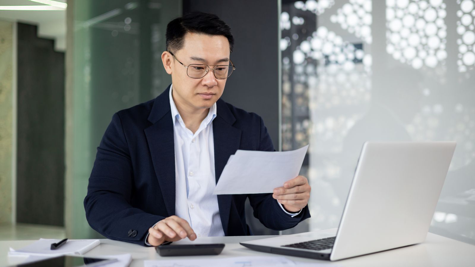 Man in glasses reading a document beside a laptop and calculator
