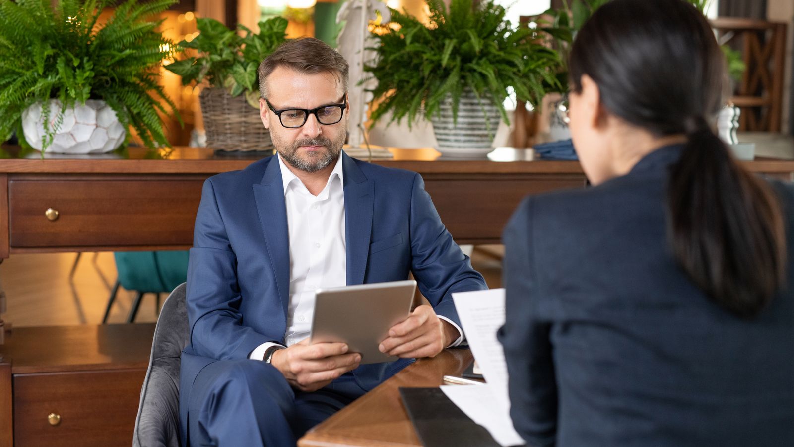Man in a suit reading a tablet across a desk from a woman holding papers