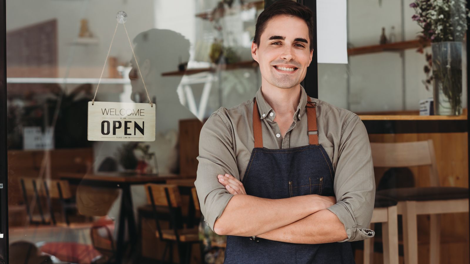 Smiling man in an apron with arms crossed outside a cafe with an Open sign