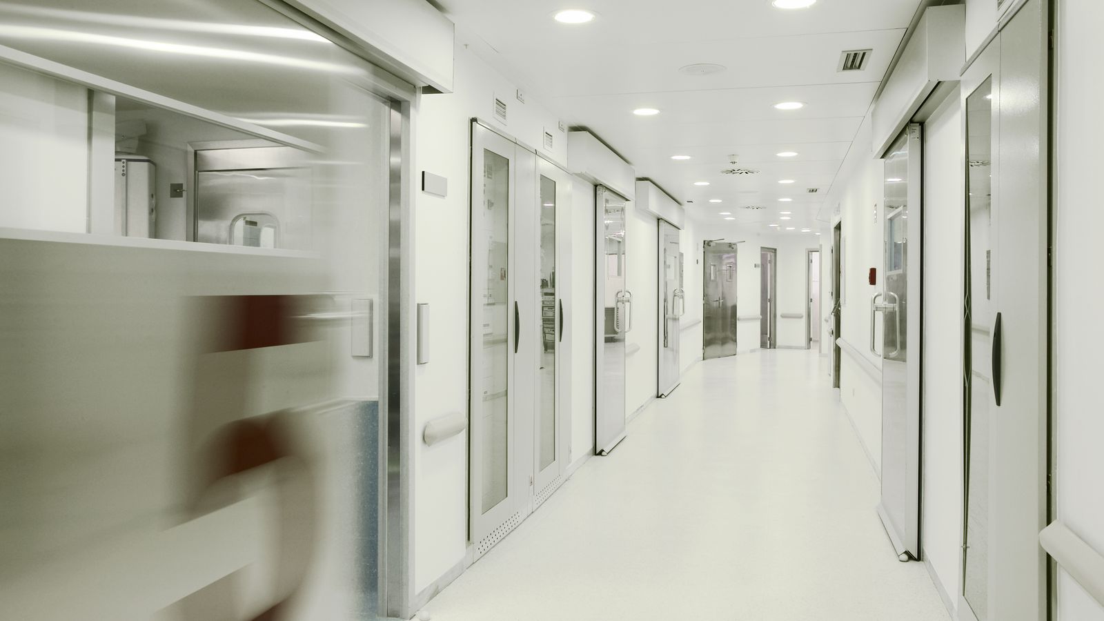 Bright hospital corridor with glass doors and a blurred figure passing