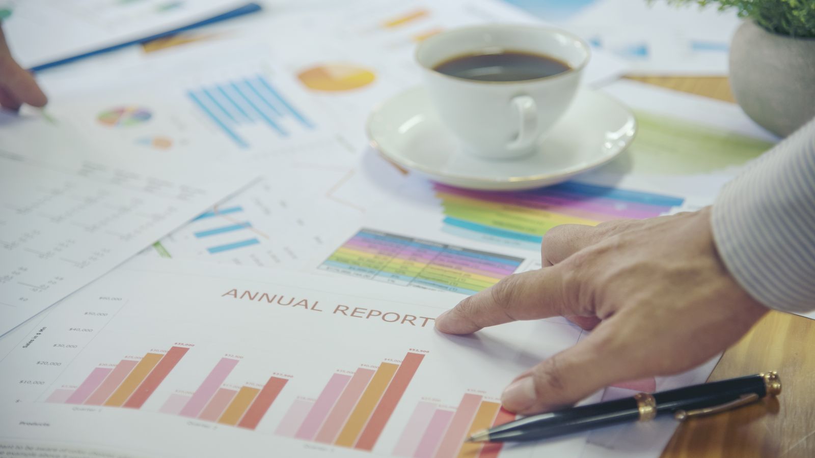Hand pointing at bar charts on a printed annual report, with a pen and a cup of coffee
