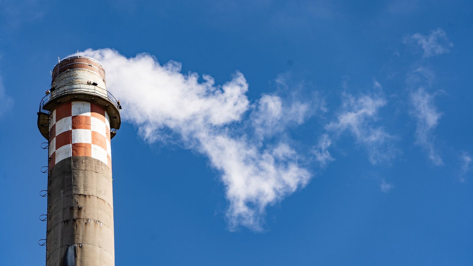 Tall concrete industrial chimney with red and white bands releasing a white plume against blue sky
