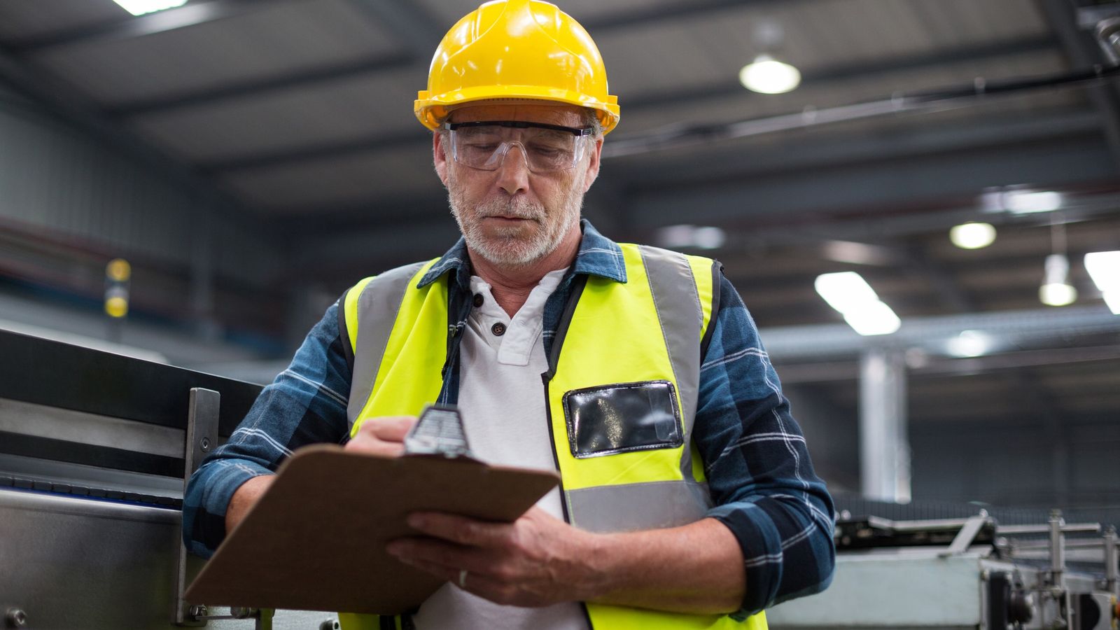 Engineer in hard hat and hi-vis writing on a clipboard beside factory machinery