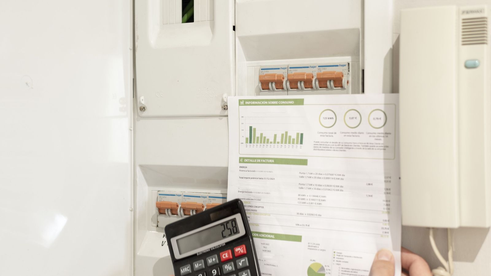 Hand holding an energy bill with usage charts and a calculator in front of a household fuse box