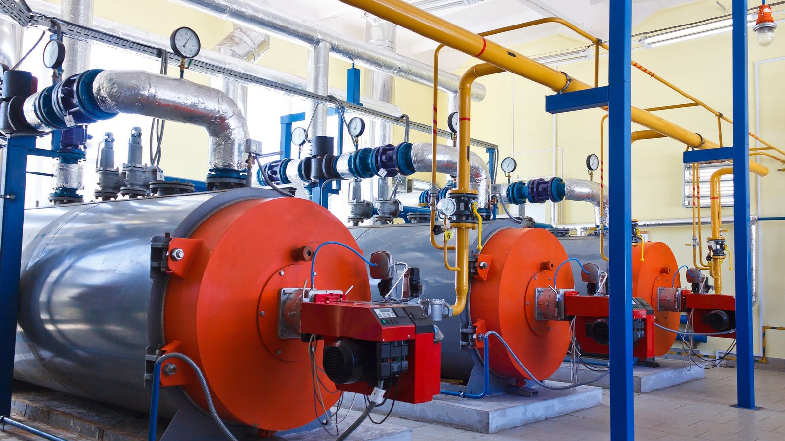 Row of large industrial boilers with orange fronts, red burners and yellow and silver pipework